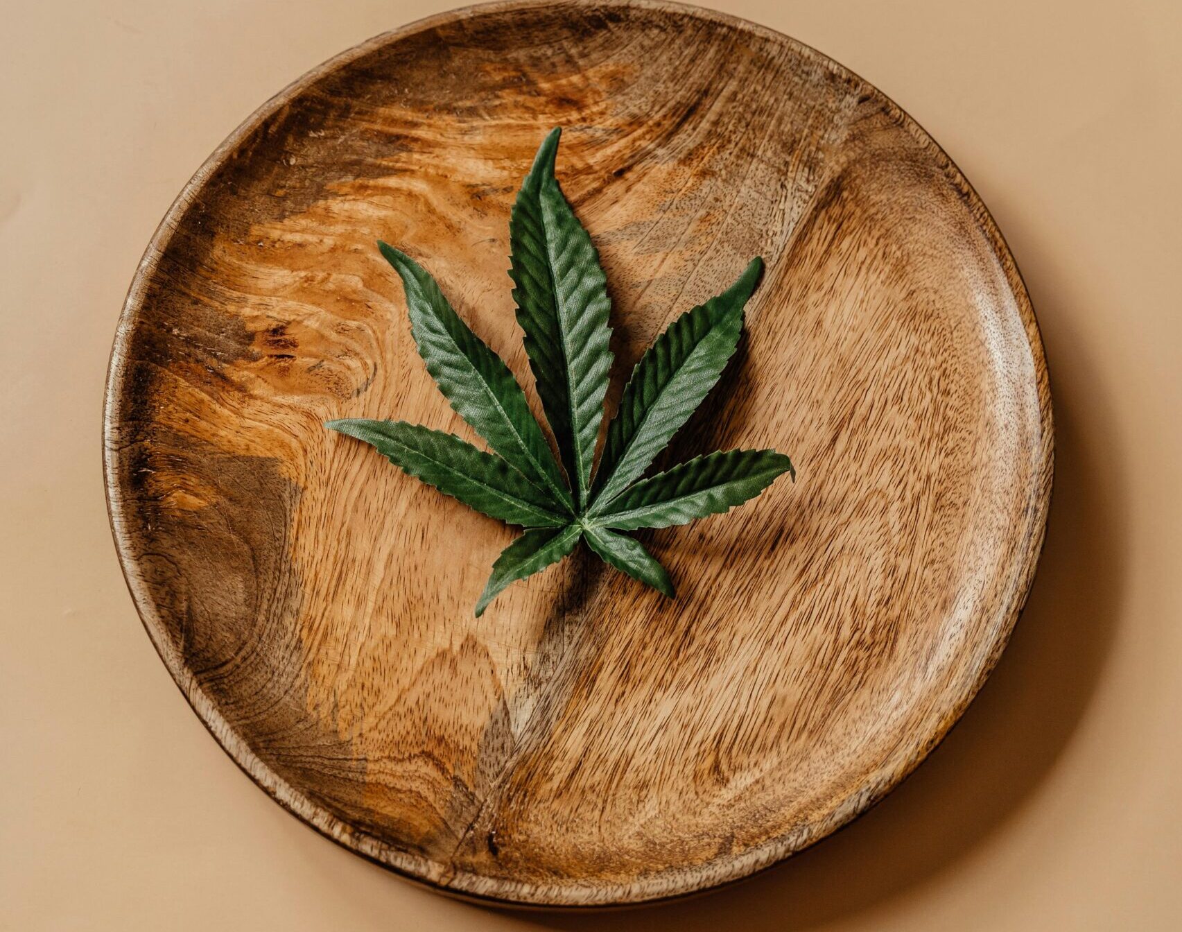 Top view of a cannabis leaf on a wooden plate against a beige background.